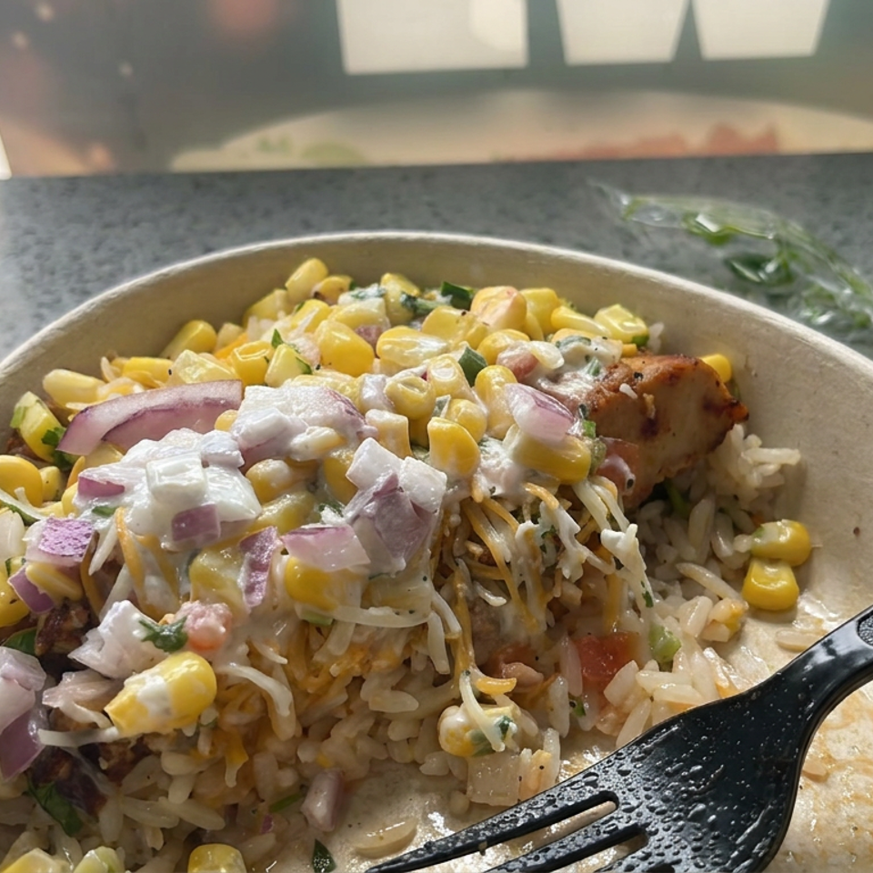 Bowl of rice topped with grilled chicken, corn, red onions, creamy sauce, and fresh herbs on a restaurant counter.